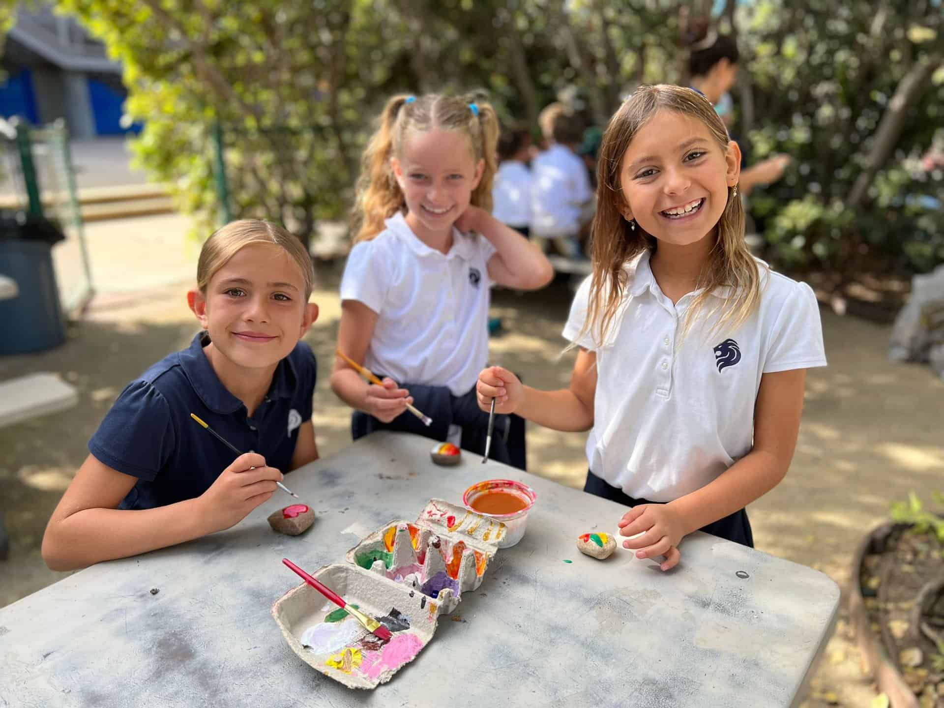 Three girls in SDJA uniforms, painting outdoors in Levana's Garden and smiling for a photo.