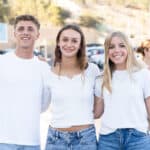 Three high school students in white tops and blue jeans smile outdoors