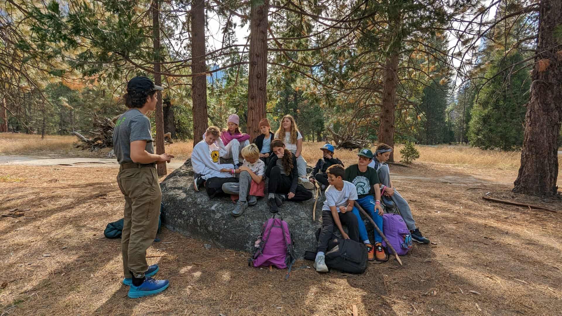 While participating in an experiential learning trip to the mountains, students sit on a rock while listening to their instructor.