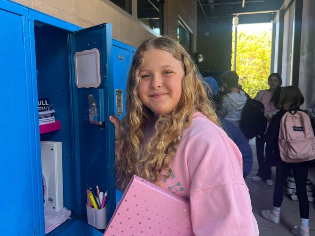 Student in pink sweatshirt gathering belongings from locker in outdoor hallway at SDJA.