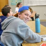 Boy with a backwards baseball cap smiles at his desk.