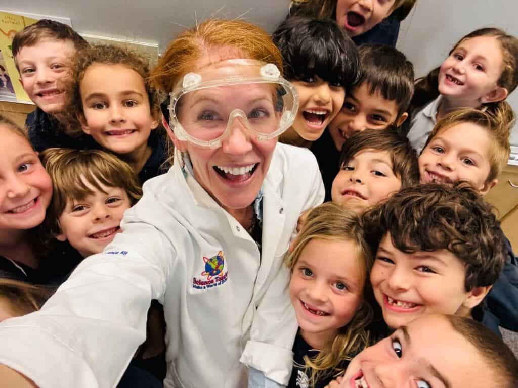 A teacher wearing a lab coat and goggles takes a selfie with her students