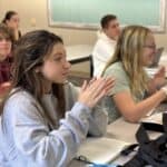 Students clap their hands while sitting at desks in a classroom.