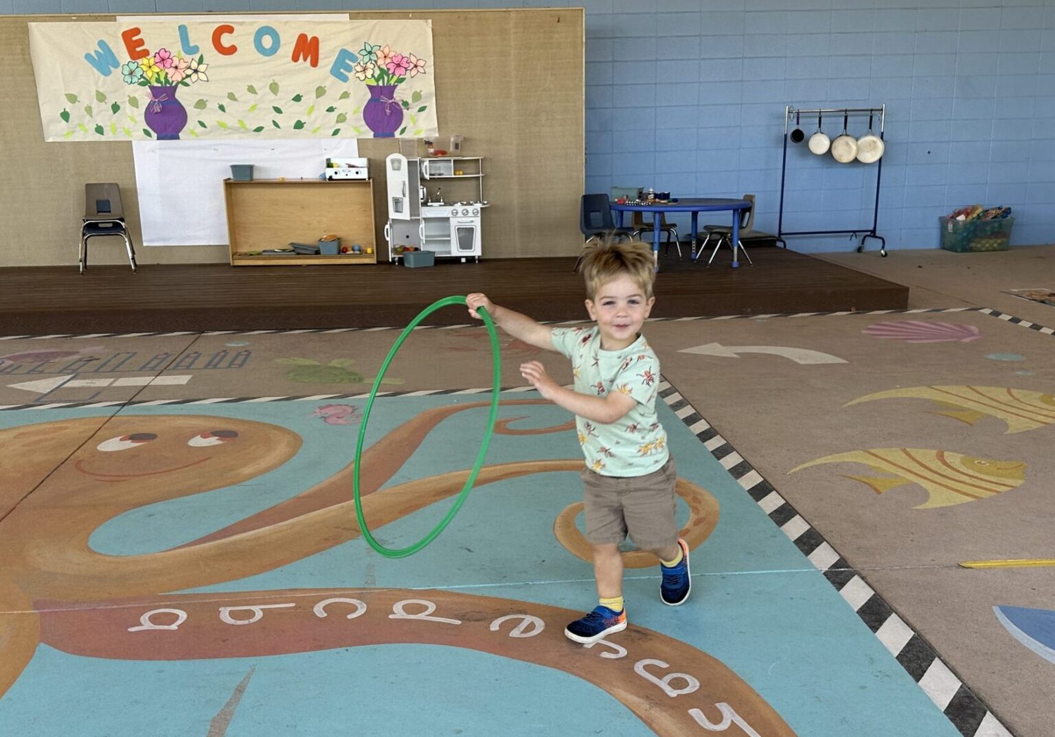 A preschooler runs with a large green hula hoop in his hand.