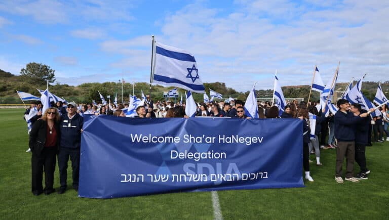 Zvi Weiss, head of school at San Diego Jewish Academy, and students from SDJA and Sha’ar HaNegev Regional High School stand in front of a banner and hold Israeli flags