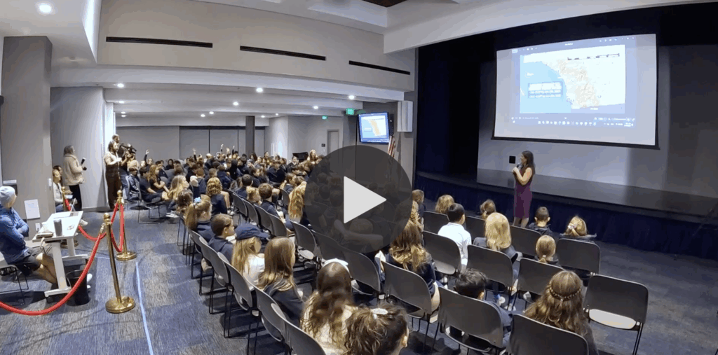 video still of SDJA class listening to a presentation in an auditorium