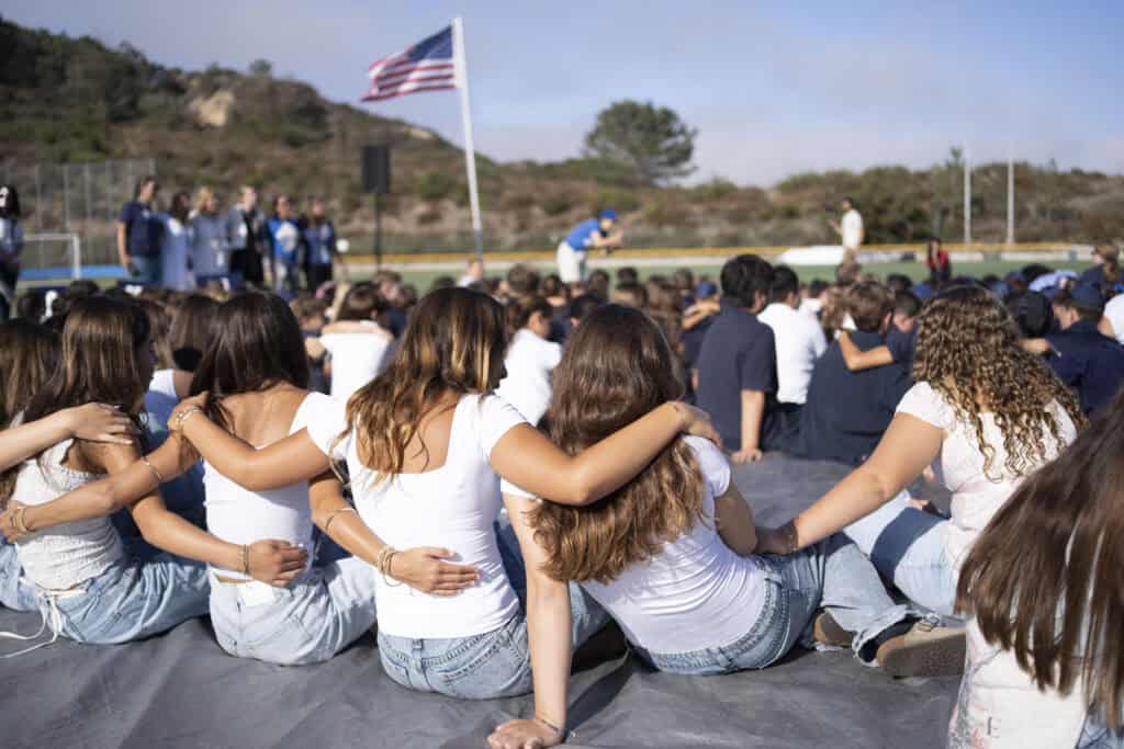 SDJA middle school girls with arms around each other, sitting on a field during a welcoming event.