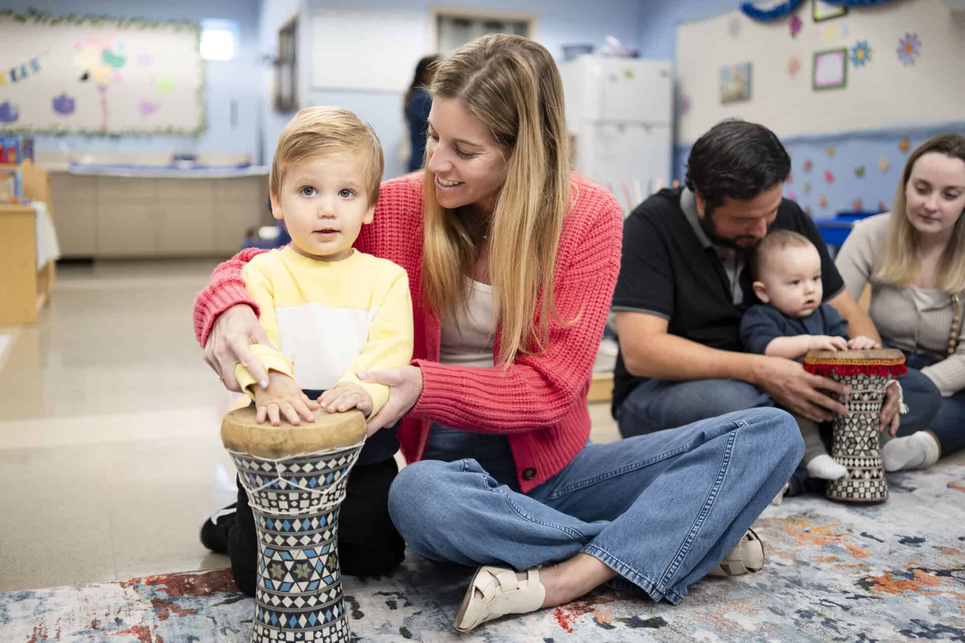 A teacher sitting next to a preschooler, helping him play a drum.