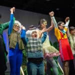 Students in costume, holding up their hands, during a performance of Shrek The Musical
