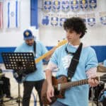 Student in light blue SDJA T-shirt playing guitar during Tikkun Olam Day performance in the gym. \