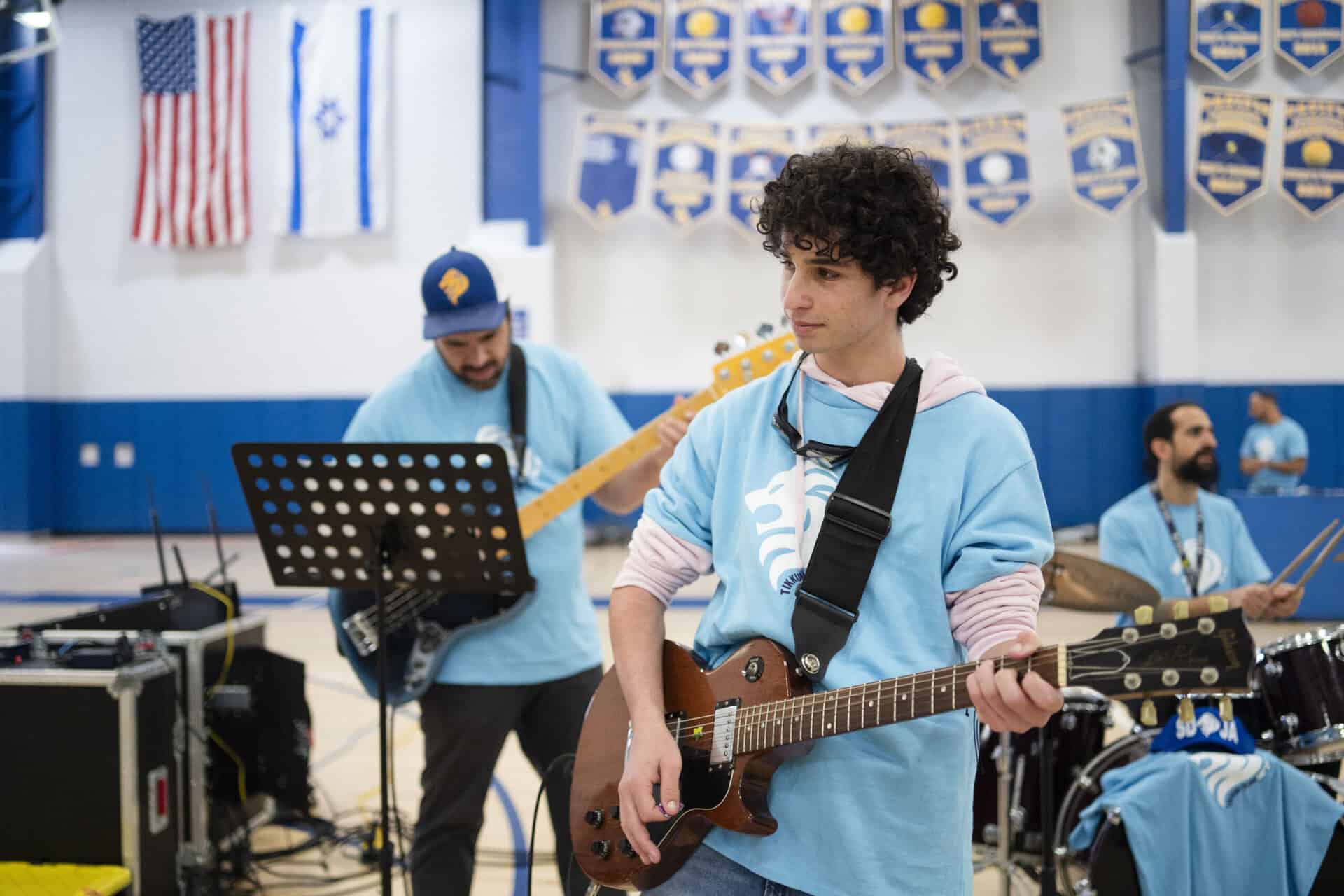 Student in light blue SDJA T-shirt playing guitar during Tikkun Olam Day performance in the gym. \