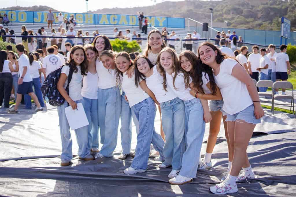 A group of young people in white tops and jeans pose together on a tarp at an outdoor event, with spectators and a "SDJA JEWISH ACADEMY" banner visible in the background.