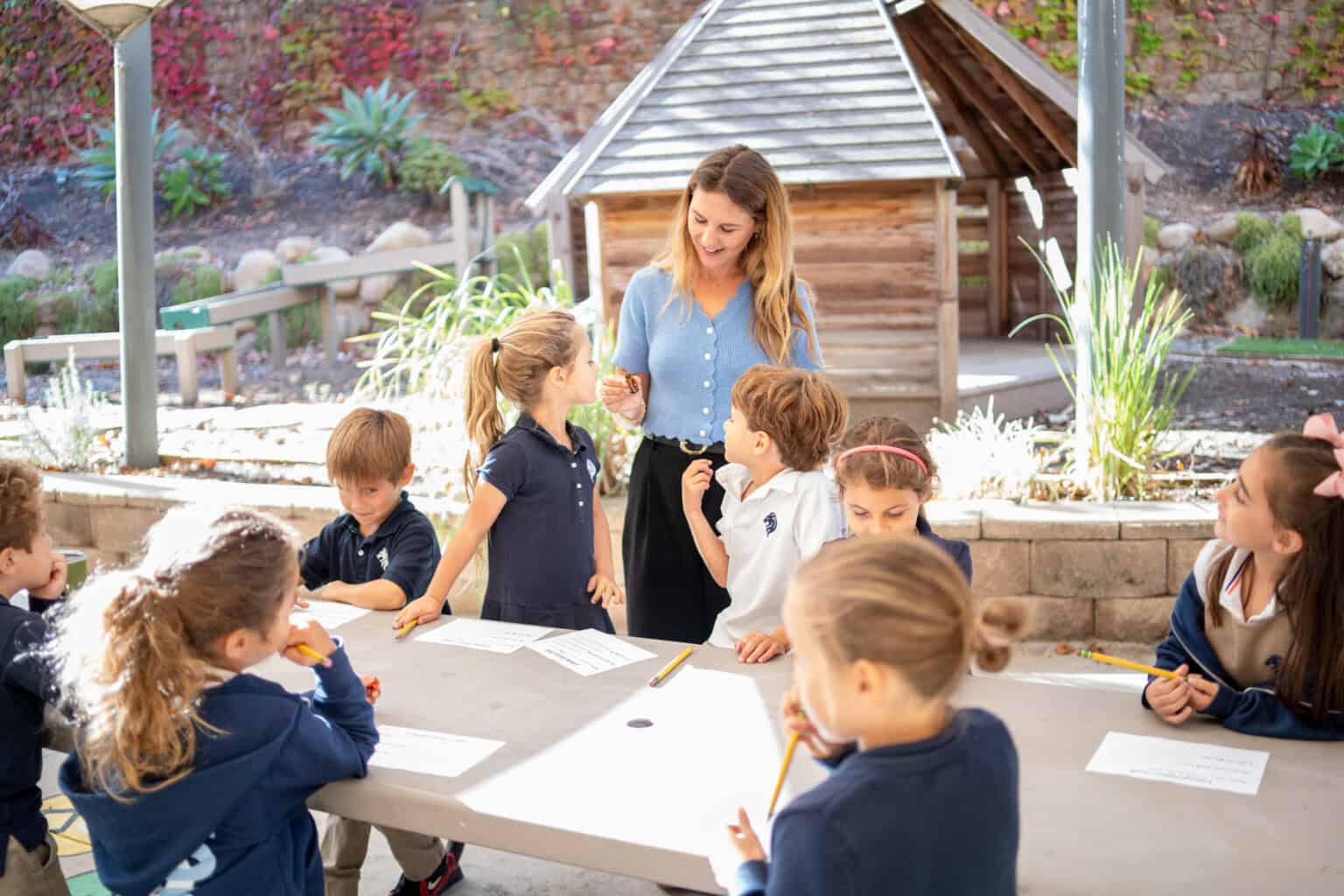 A teacher guides a group of young students as they work together at a table outdoors in a garden setting.