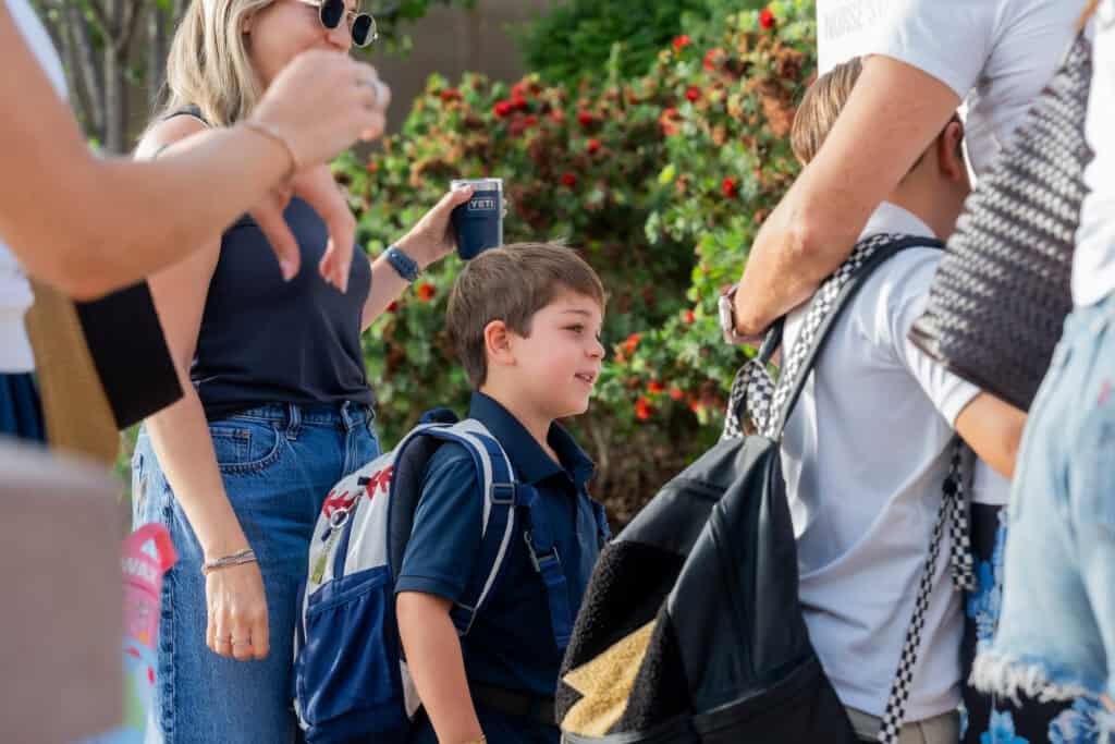 A young child with a backpack stands among adults in an outdoor setting, holding a drink container.