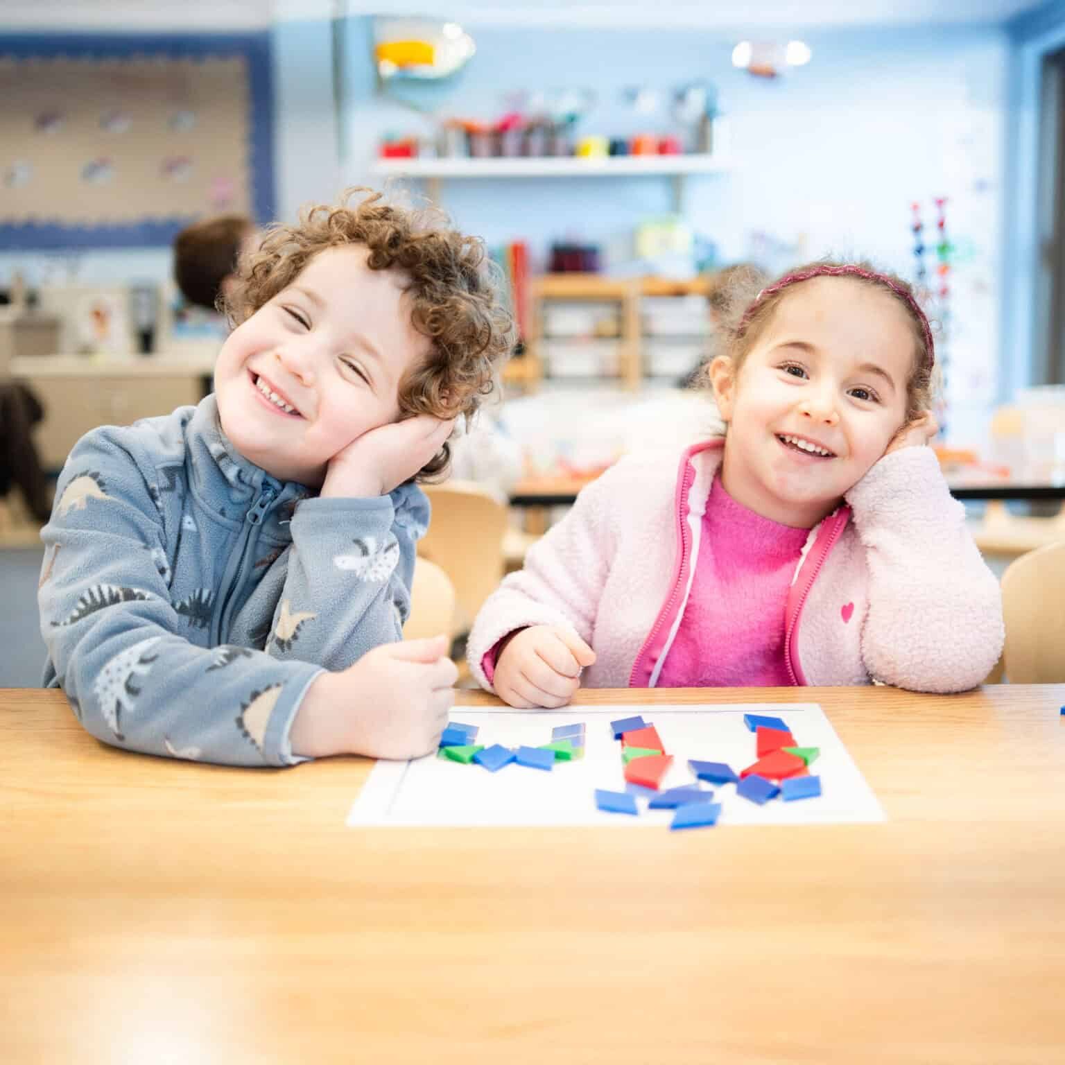 A pair of preschool students smiling, while seated at a table playing with blocks.