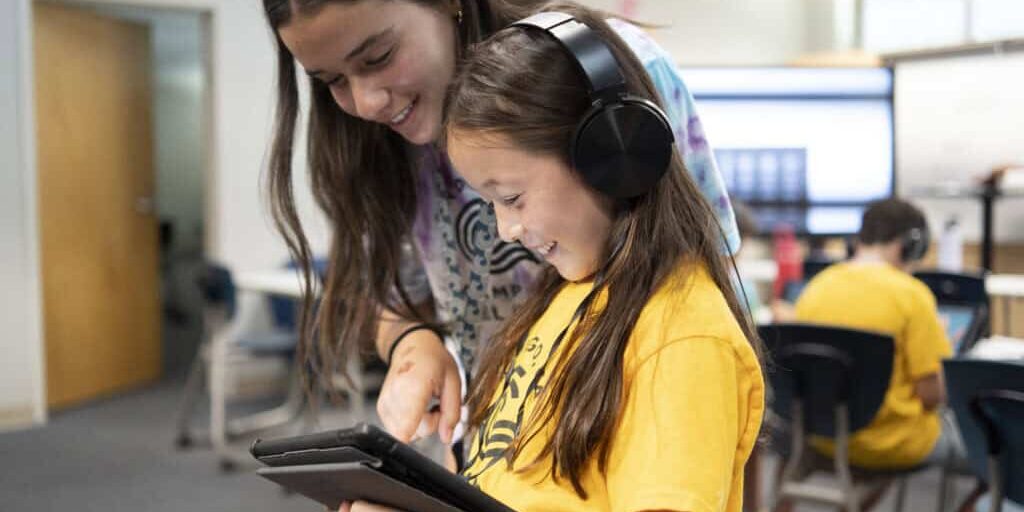 A girl using a tablet with headphones standing next to a camp counselor.
