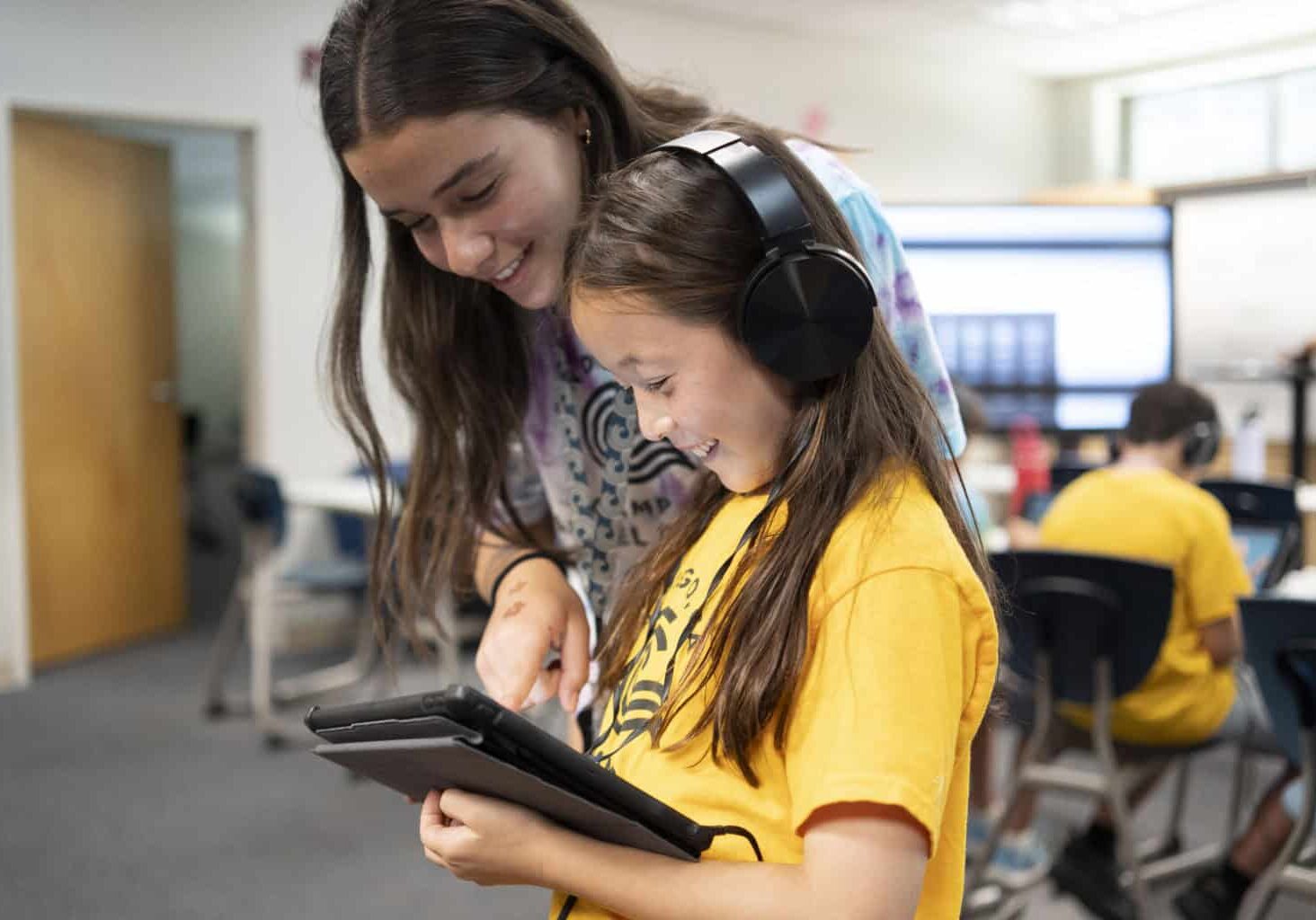 A girl using a tablet with headphones standing next to a camp counselor.