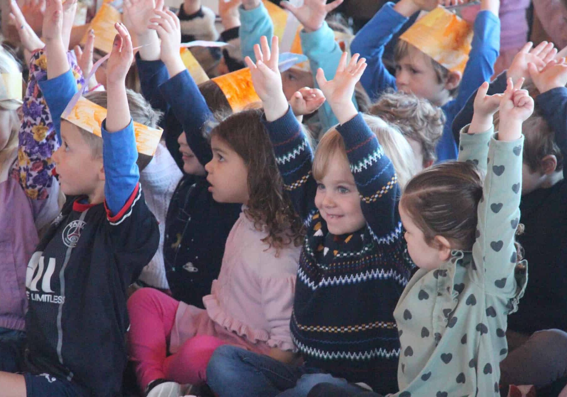 Preschool students sit on the floor of a multipurpose room with their hands raised in the air.