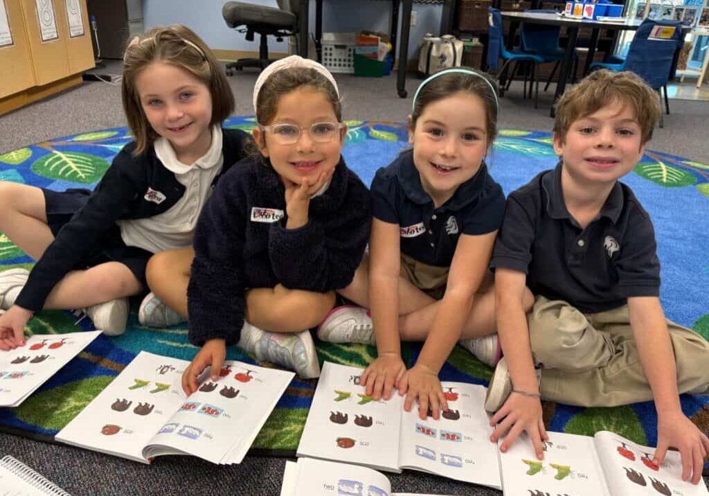 a group of elementary school students sit together on a rug and read from their workbooks.