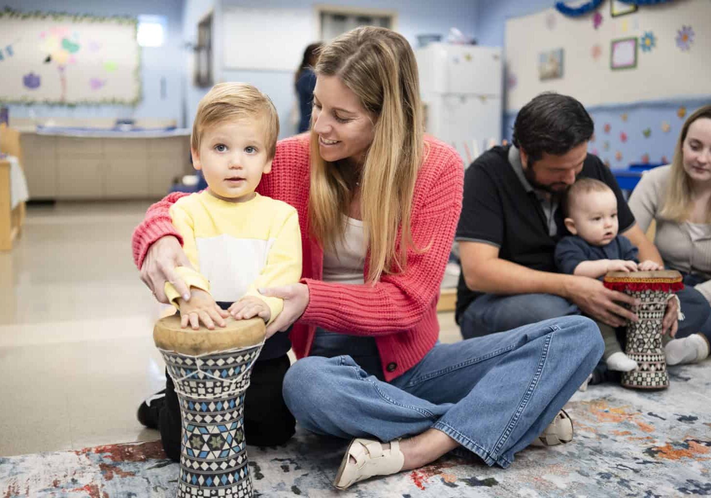 A teacher sitting next to a preschooler, helping him play a drum.